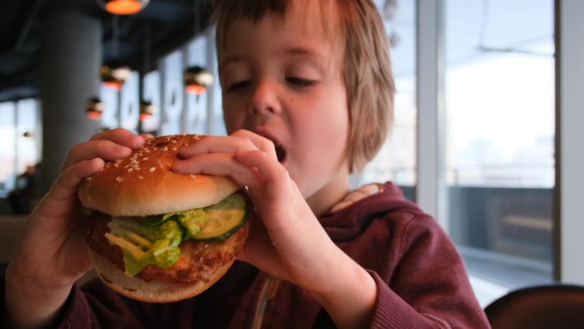 Little boy in fast food cafe eats burger. Portrait of hungry child Cute little kid eating burger. Hungry child in fast food cafe. Close-up boy eating burger. Hungry child in cafe. Lovely boy