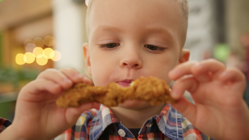 Portrait hungry blond 4-year-old boy sitting fast food and eating fried chicken in breadcrumbs. Caucasian kid satisfies his hunger eating deep-fried chicken from fast food. Junk food from fast food.