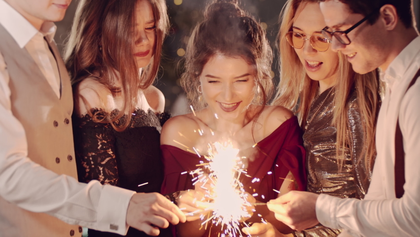 Beautiful friends holding christmas sparklers in hands. Guys celebrating by waving fireworks, enjoying party on New Years or Christmas Eve night. Happy group of people enjoying party in Xmas evening.