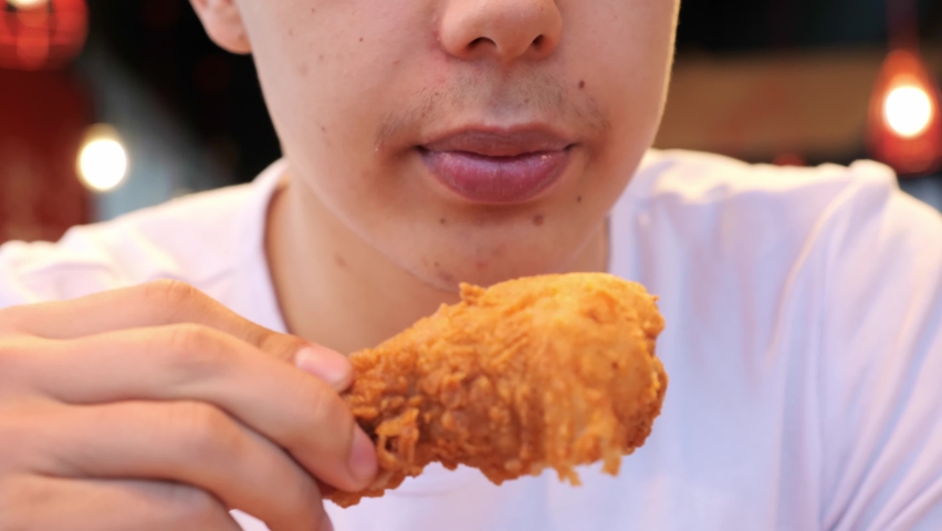 Handsome young man student eating chicken wings, high calorie food, health risks, cholesterol. Close up of male mouth eating junk food indoors. The guy bites a crispy chicken fried in oil.