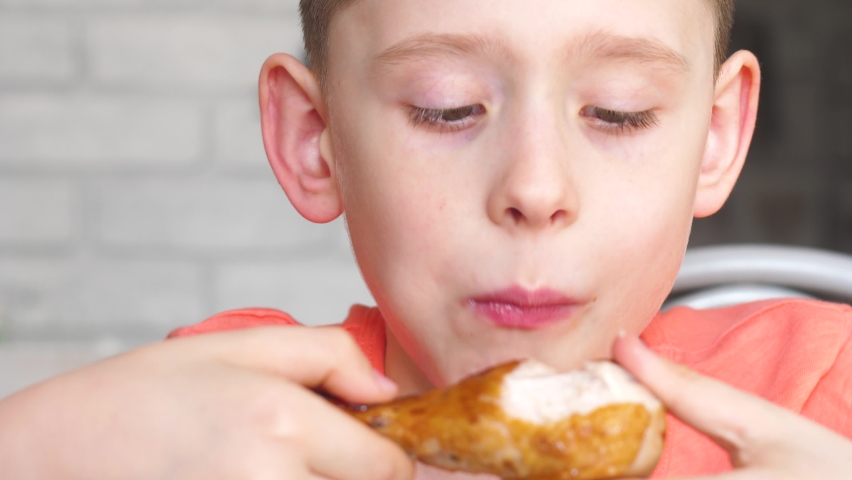 Portrait of a caucasian boy 8-9 years old eating fried chicken legs, closing his eyes in pleasure. the child eats meat with his hands in a restaurant. fried chicken lunch