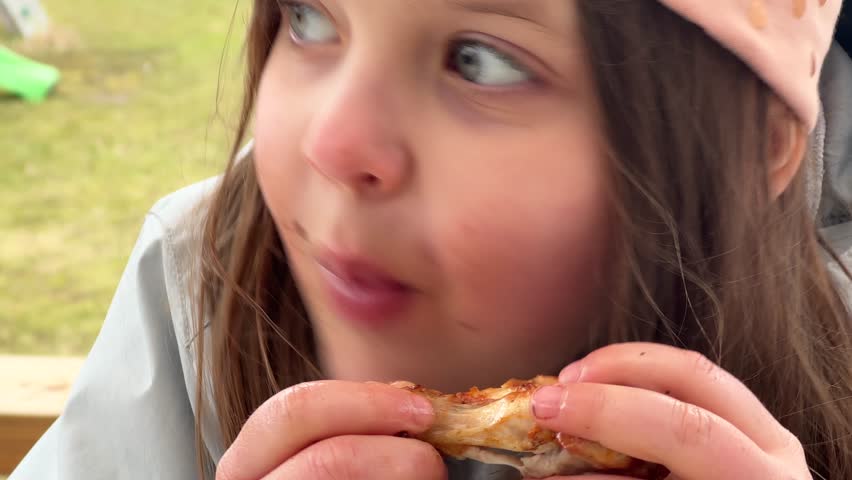 Close-up of a young girl with blue eyes, eating a chicken wing with an eager expression, outdoors, with long hair and a cap.