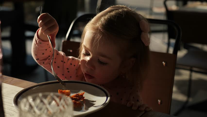 Adorable little girl eating meal using fork at the restaurant. 2 years old kid skills