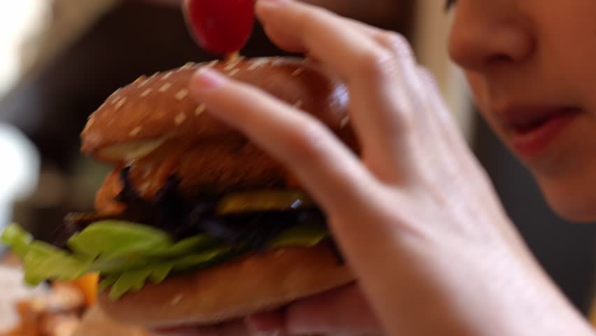 Boy eating crispy chicken burger with lettuce, pickles, and tomato in restaurant. Close up shot of child biting hamburger. Hungry boy eating unhealthy food