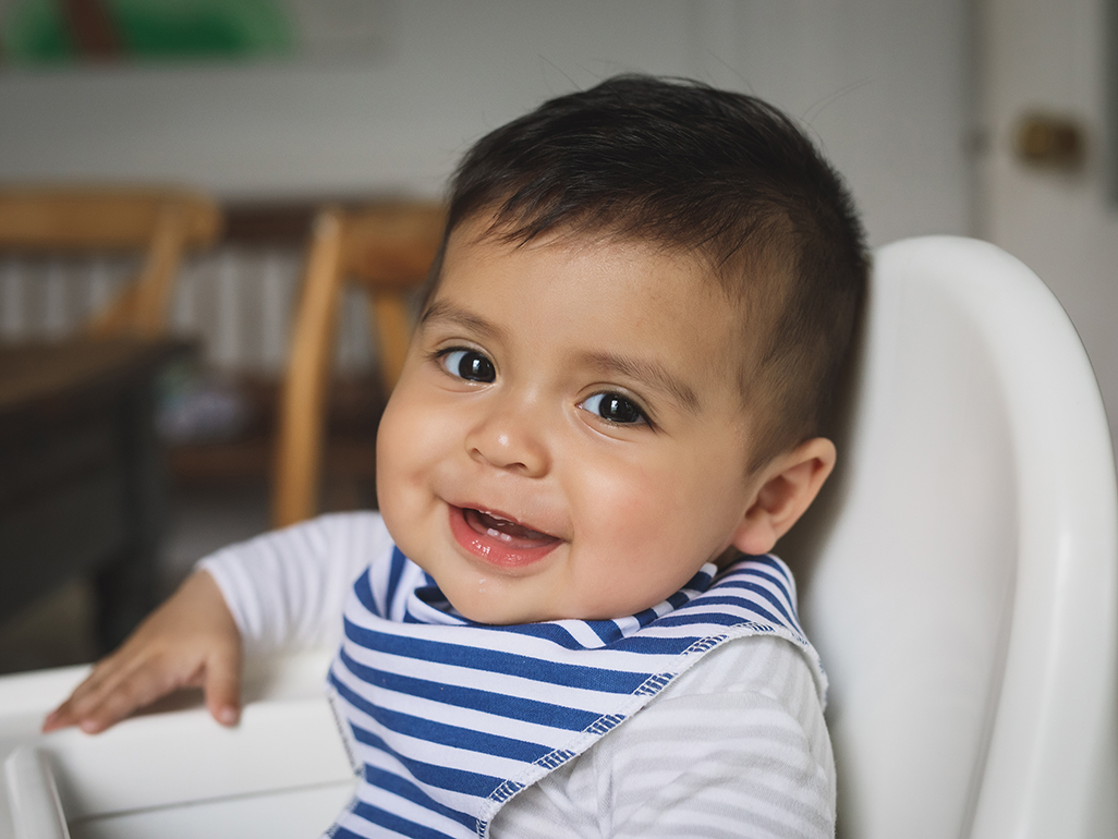 Hispanic baby boy sits in high chair and smiles.