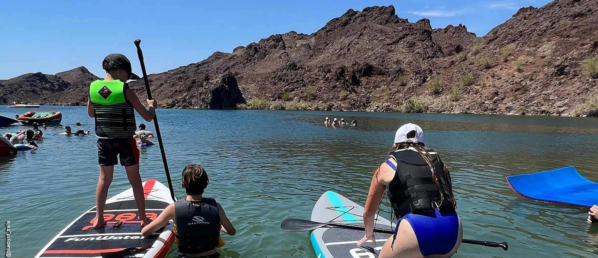 A kid standing on a paddleboard and another sitting on a paddleboard with one child in the water