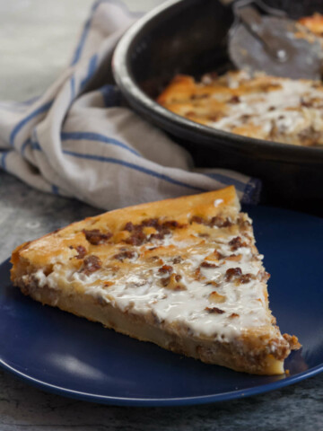 Piece of pie on a blue plate, a napkin and a pan with pie in the back, on a gray background.