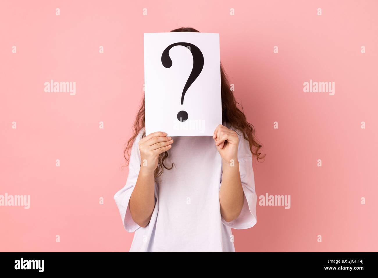 Portrait of anonymous little girl wearing white T-shirt hiding her face behind white paper with question mark, finding smart solution, asking for advice. Indoor studio shot isolated on pink background Stock Photo