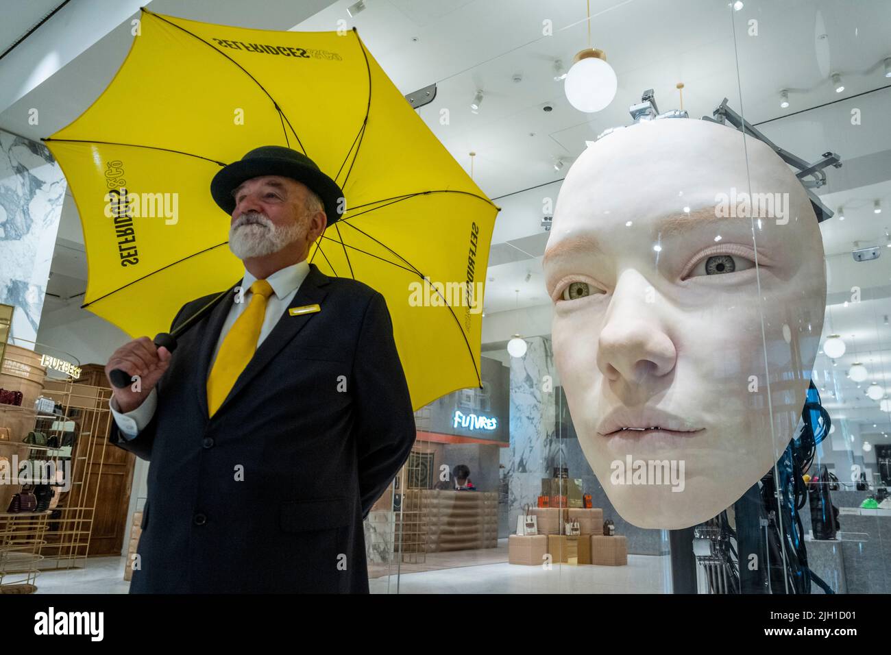 London, UK.  14 July 2022. A Selfridges doorman poses with 'The Giant', 2021, by Gentle Monster, a nine foot tall face with hyper realistic movements and expressions at a preview of SUPERFUTURES, a new exhibition at Selfridges, Oxford Street.  13 major large-scale immersive art works on display across the store and windows present contemporary artists’ visions for the futures.  The exhibition is part of Selfridges’ initiative called ‘reinventing retail’ and runs to 15 October 2022. Credit: Stephen Chung / Alamy Live News Stock Photo