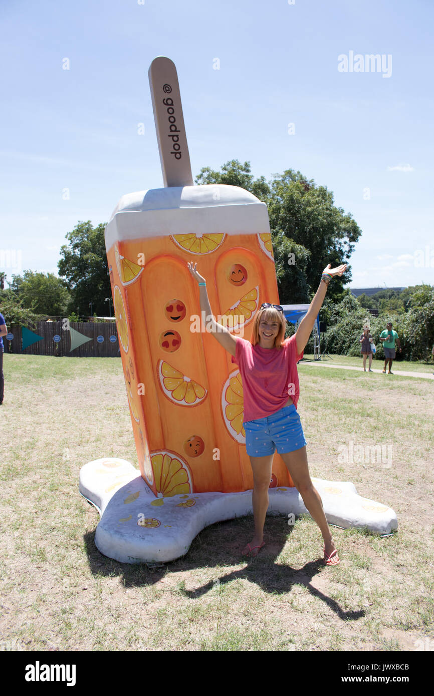 Young blond woman with pink shirt and blue shorts arms raised smiling in front of large orange Popsicle model outdoors at Ice Cream Festival. Stock Photo