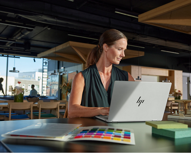 A woman with a ponytail sits at a table in a modern office space, working on an HP laptop