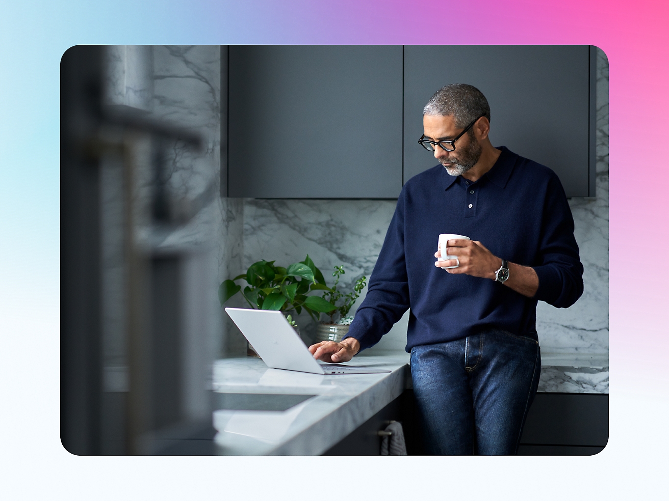 A man stands in a kitchen, holding a mug and looking at a laptop screen on the counter.