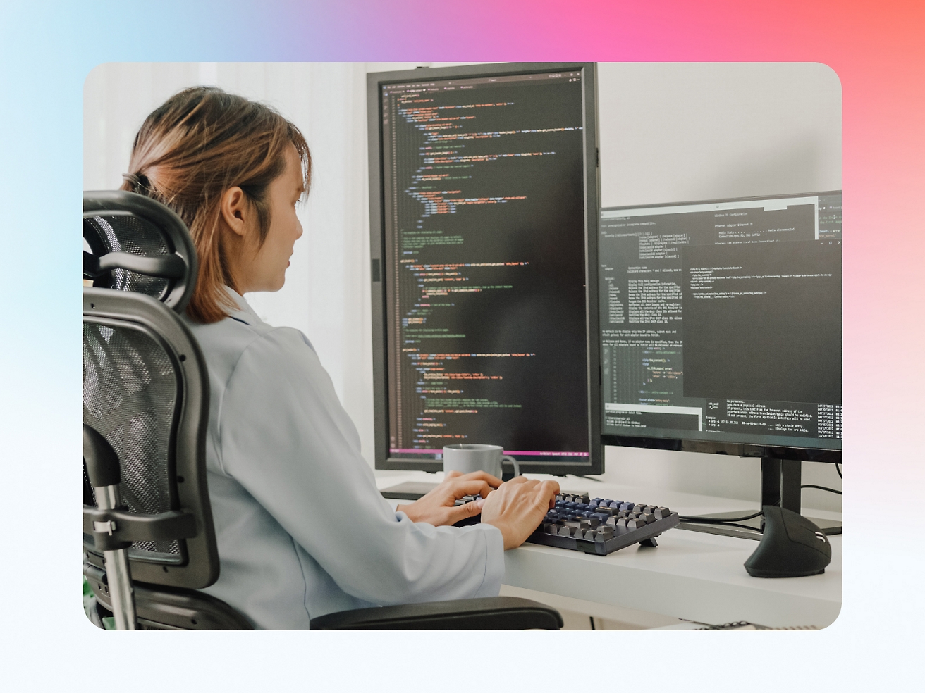 A woman sitting in an office chair, coding on a computer with multiple screen displays showing programming code.