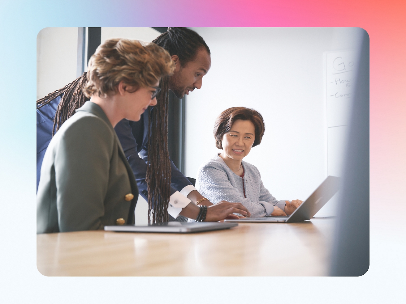 Three professionals, one with dreadlocks, sitting around a laptop in a meeting room, discussing work contentedly.