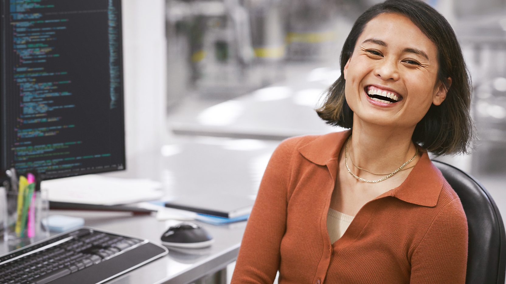 A joyful woman sitting at a desk with a computer, displaying code on the screen, in a modern office setting.