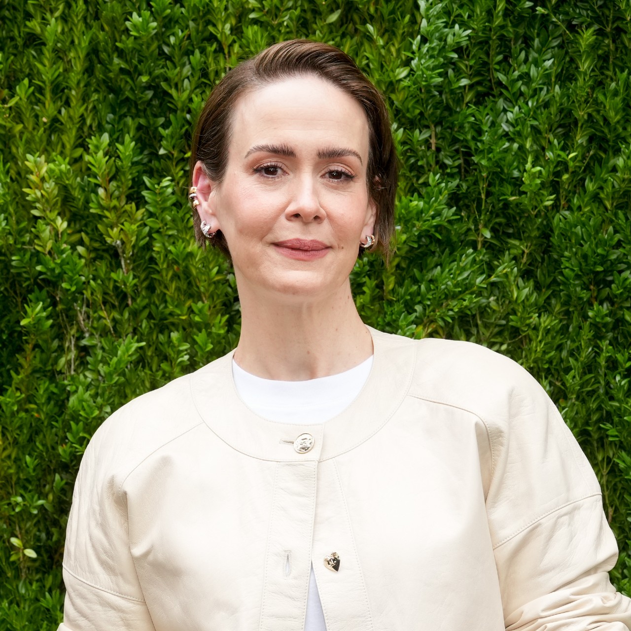 a white woman with short hair wearing a cream sweater and posing in front of a grass wall