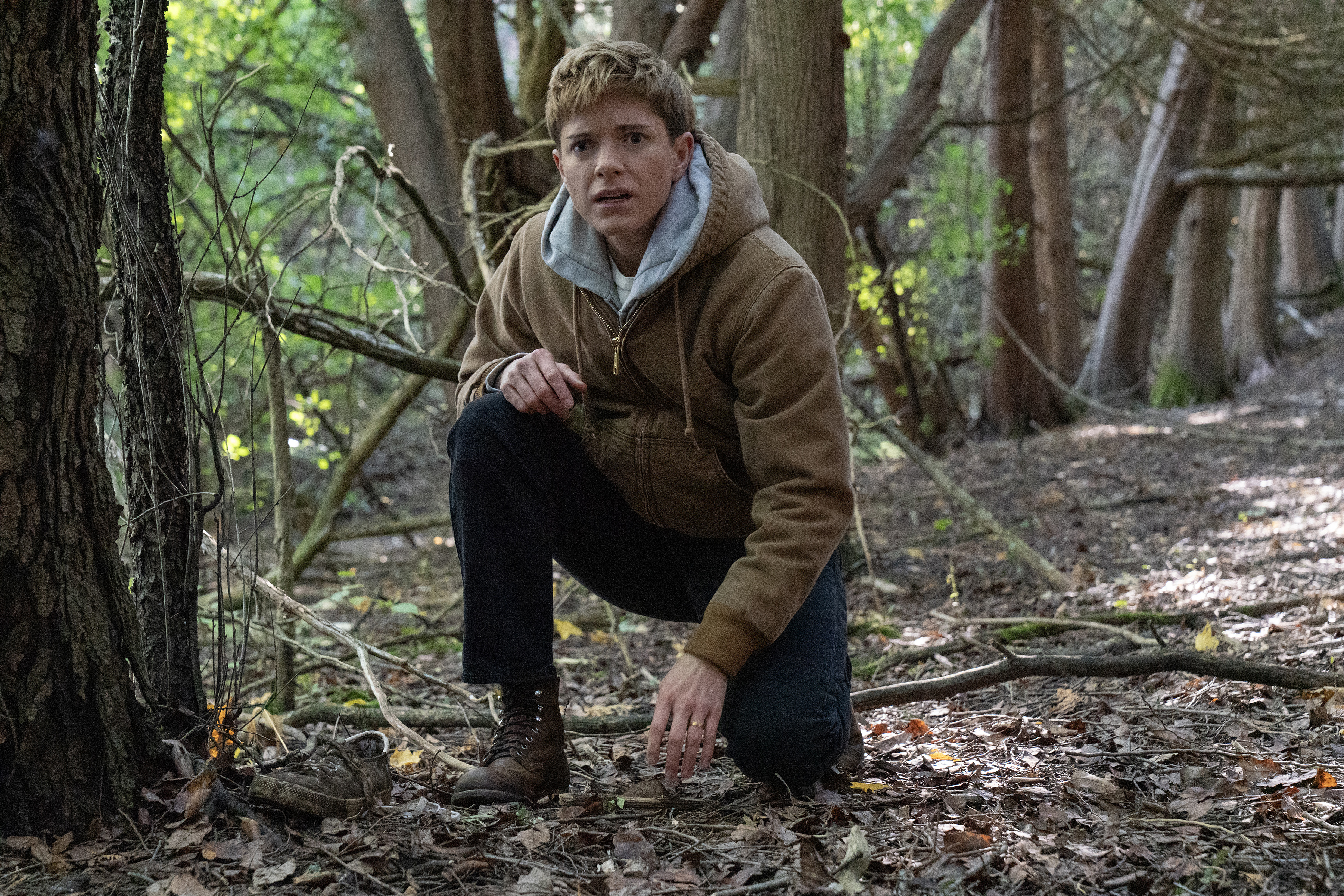 mae martin wearing a hoodie kneeling in the woods in wayward