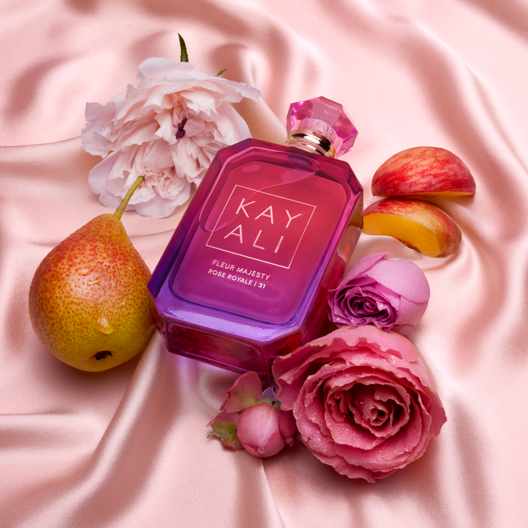 kayali fleur majesty rose royale perfume bottle laying on pink satin fabric with roses, peach slices, and pears