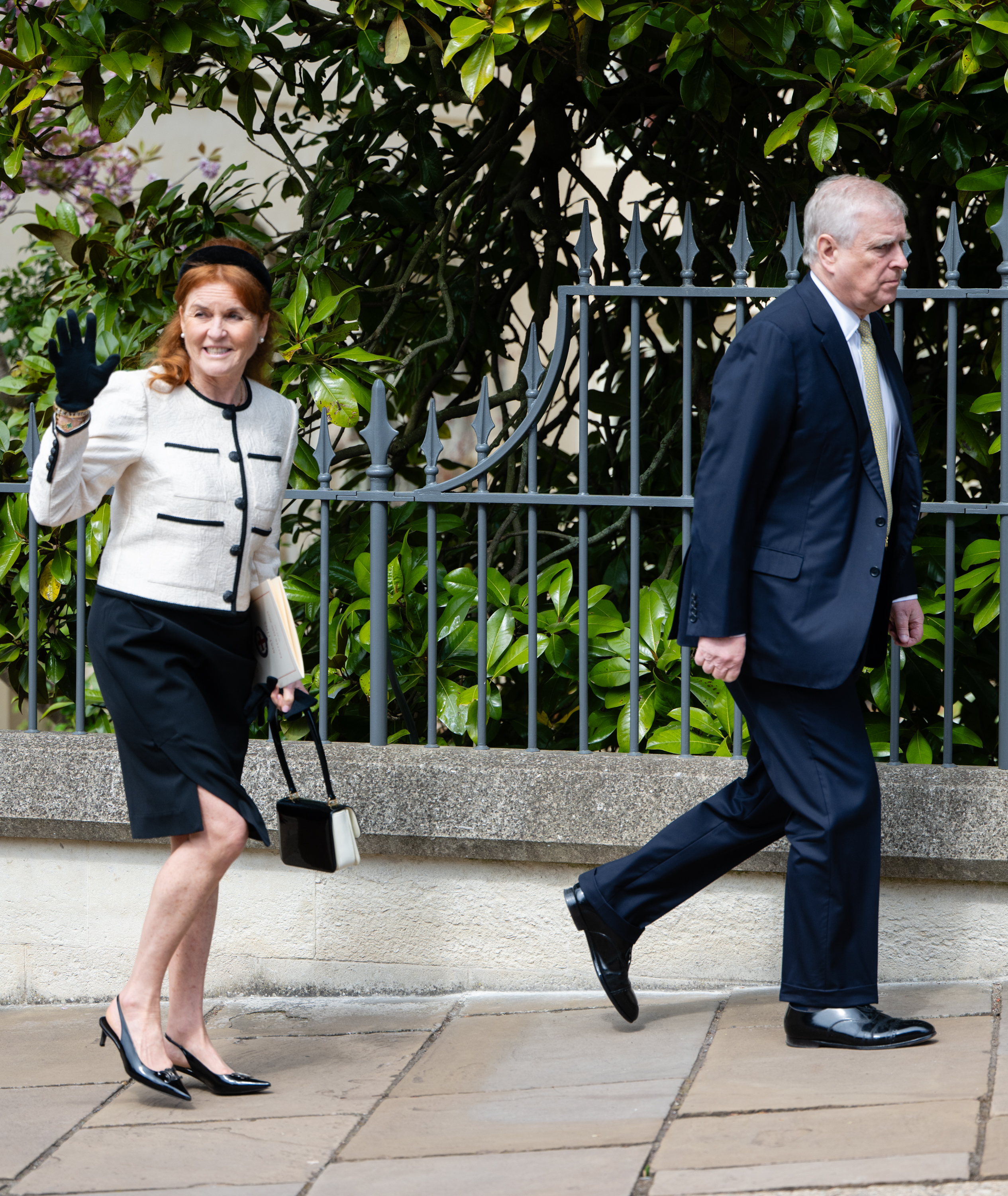 Sarah Ferguson wearing a white jacket and black skirt and waving, walking behind Prince Andrew along a fence and trees