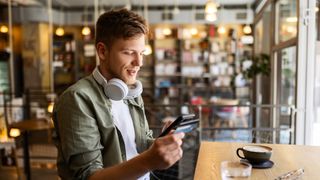 A man sits in a coffee shop, headphones around his neck, as he smiles while using his phone