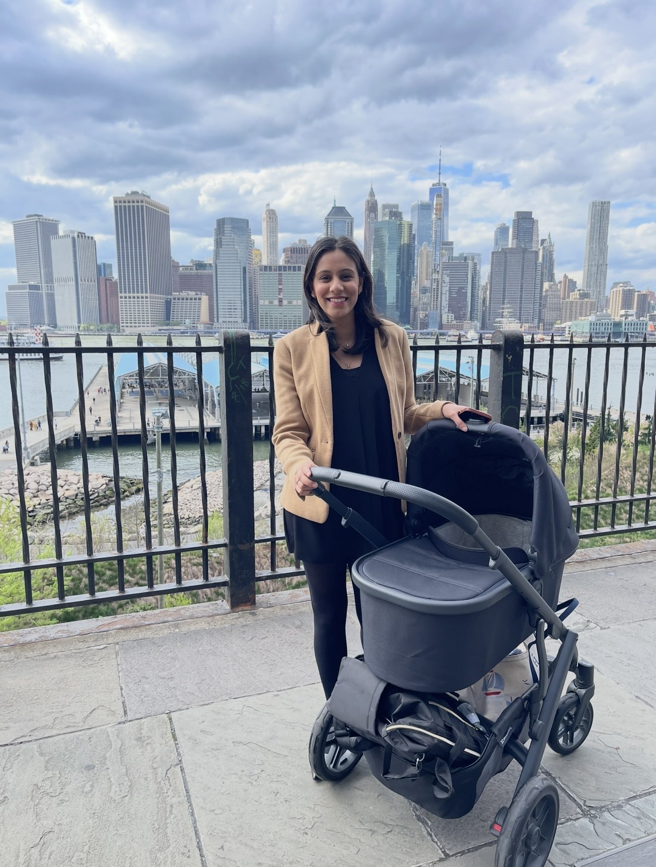 Sidra Mahfooz stands outdoors by a railing overlooking the New York City skyline, smiling while holding the handle of a baby stroller. She wears a tan blazer over a black outfit.