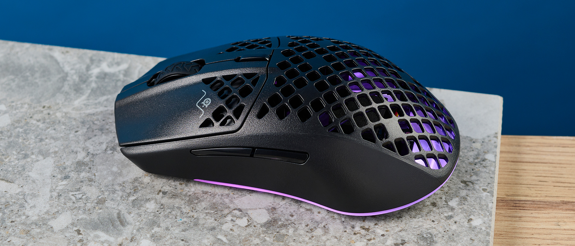 The SteelSeries Aerox 3 on a stone surface and wooden table against a blue background. 