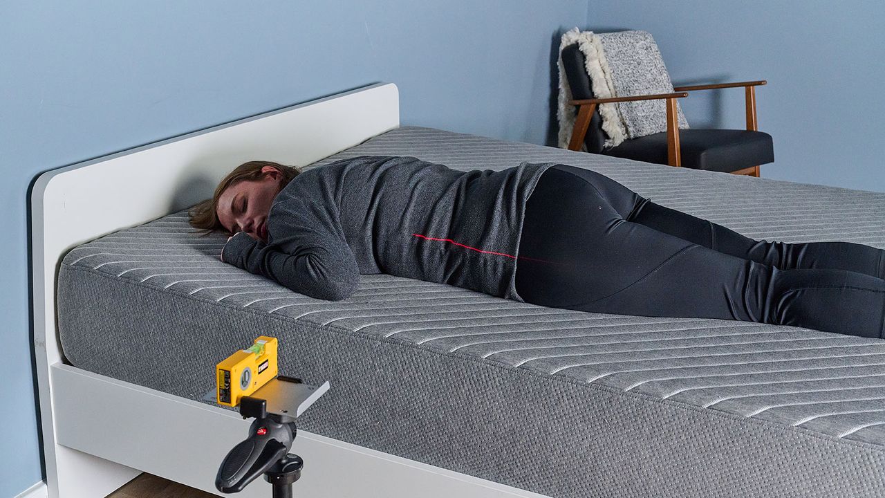 This image shows our sleep tester Ruth Jones lying on the Siena Memory Foam Mattress during the pressure relief test. She is lying on her stomach on the left hand side of the mattress, with no pillow. A laser, balanced on a tripod at the front of the shot, projects a red line on to her body. The line is positioned where her spine would be. As the body is aligned with the laser, it indicates good support for stomach sleepers.