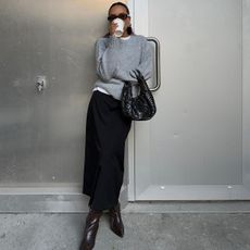 Ingrid wears a gray sweater, black woven bag, black maxi skirt, and black knee-high boots.