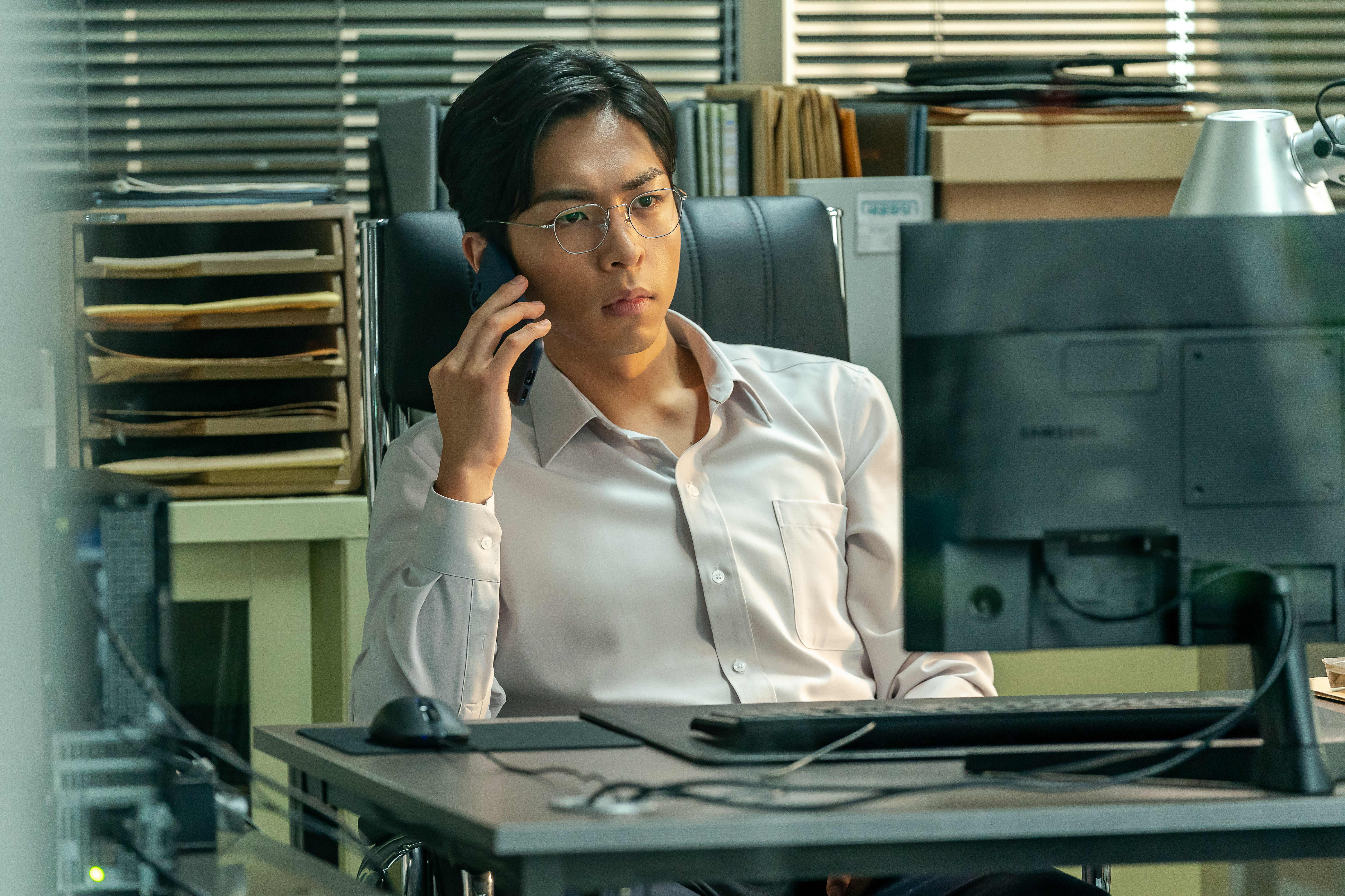 Joo Jong-hyuk sits at a desk in front of a desktop computer on the phone in 'Tempest.'