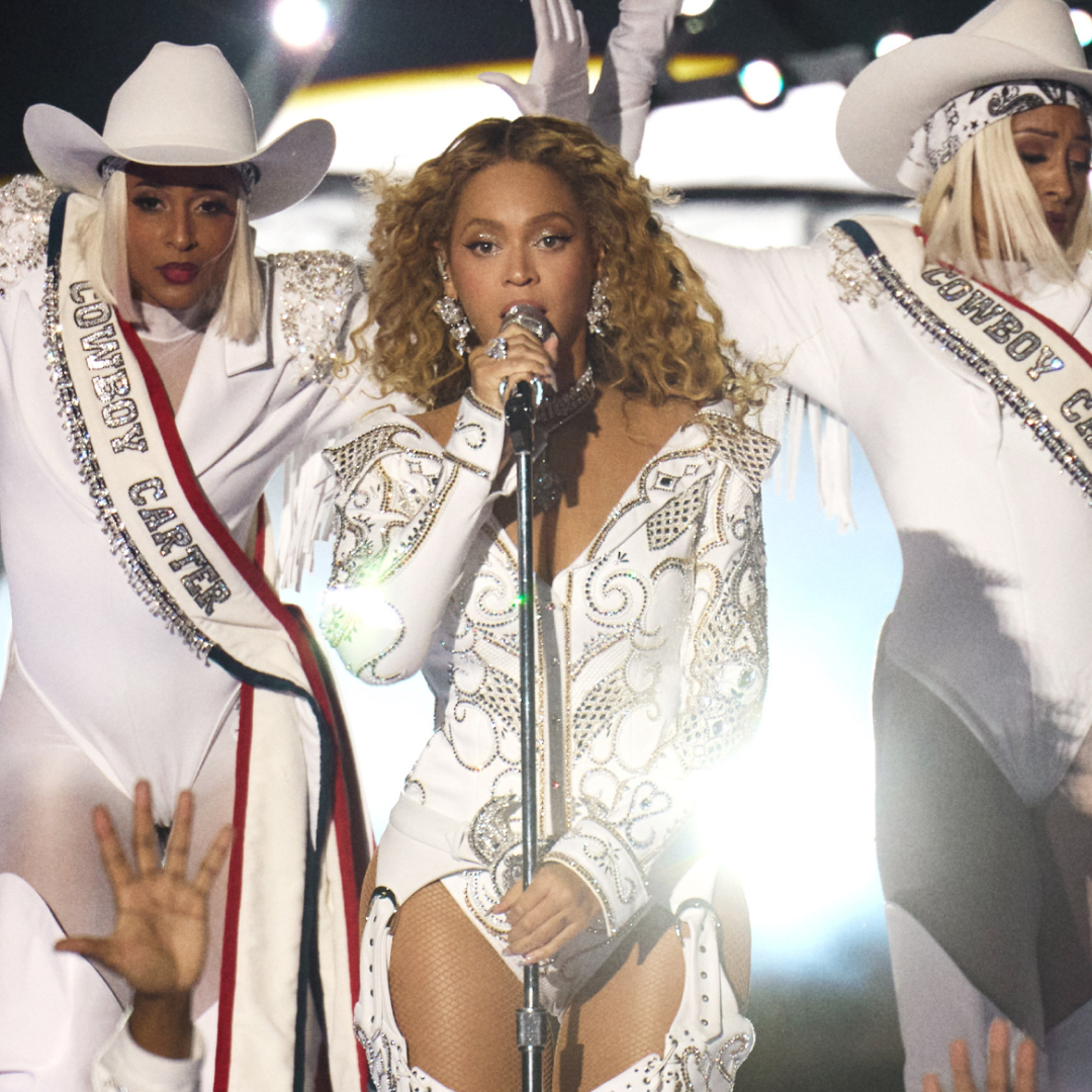 Beyoncé sings into a mic stand on stage with two backup dancers in white Western costumes, during Netflix's 'Beyoncé Bowl.'