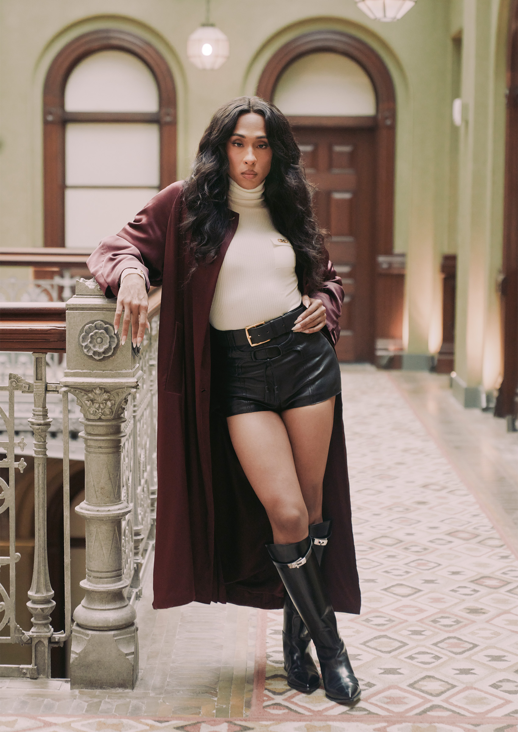 portrait michaela jae rodriguez wearing red leather jacket black shorts and black boots poses in a building hallway