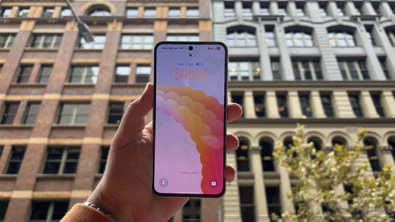 Samsung Galaxy A56 holdes op foran en h&oslash;j bygning.