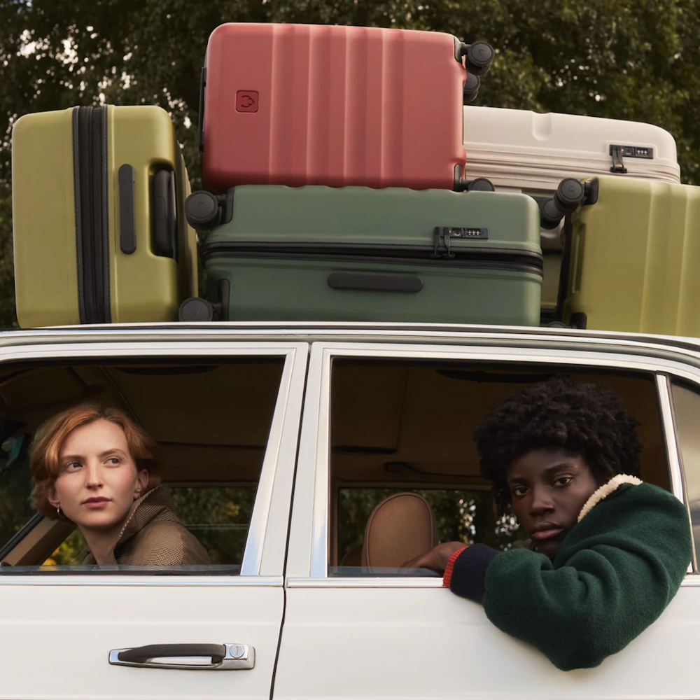 An image of the side of the car with a woman looking out of the front window and another woman hanging her arm out of the back window with several Antler suitcases on top of the car