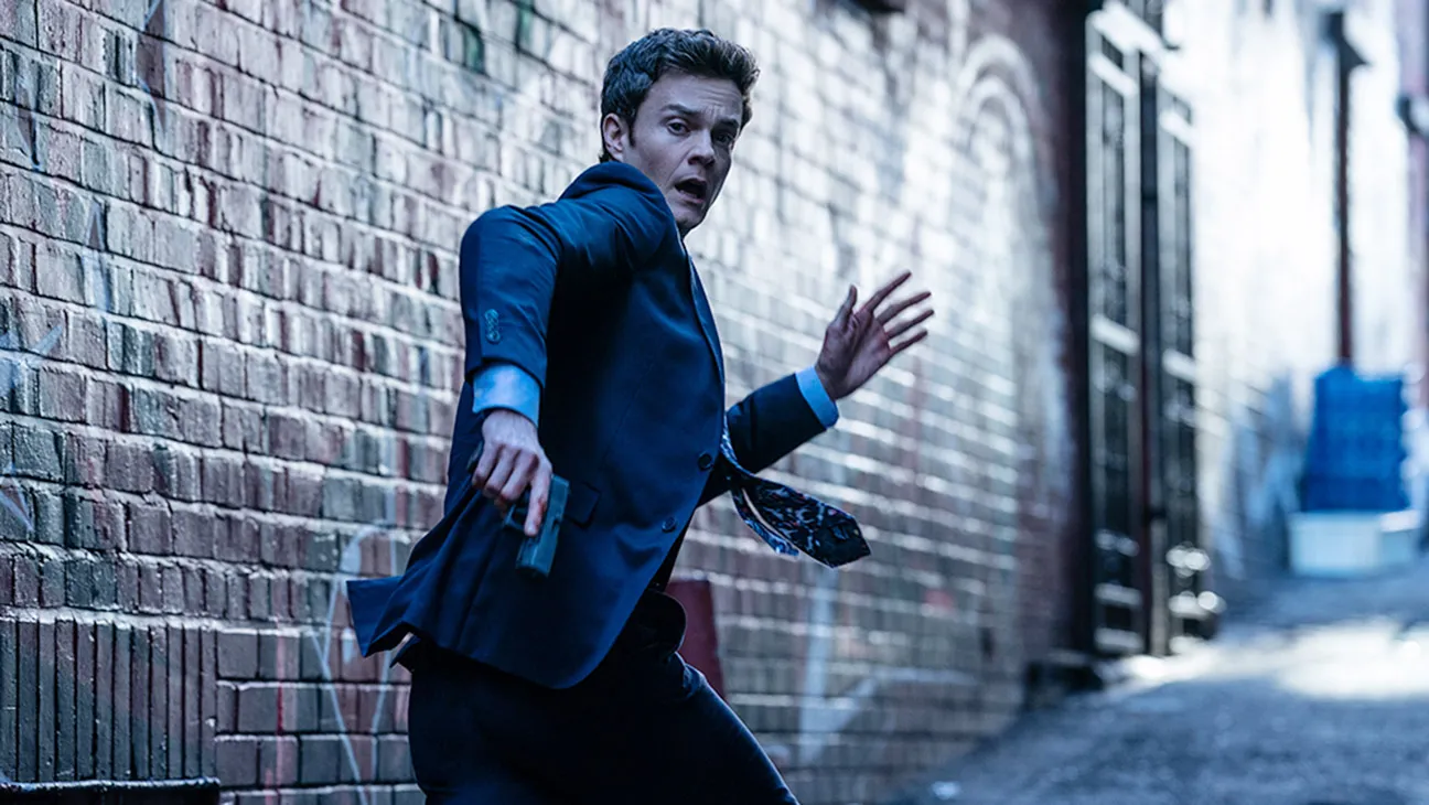 jack quiad wearing a suit and holding a gun as he runs down an alley in the movie novocaine