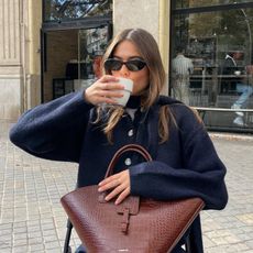 Fashion influencer  @lunaisabellaa sitting outside drinking coffee wearing a stylish knit sweater and oval sunglasses with a mock croc bag.