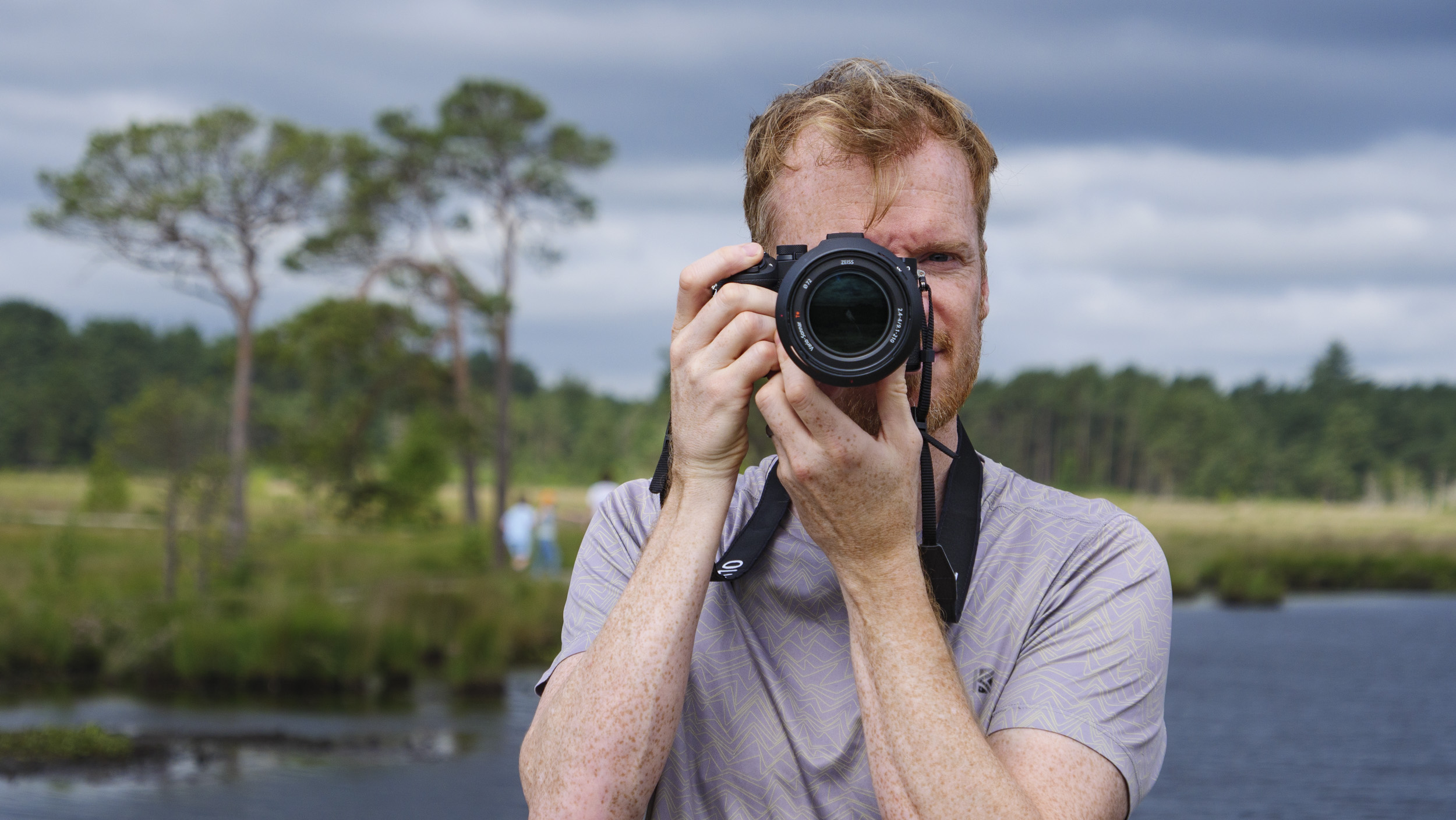 Photographer and tech journalist Tim Coleman holding the Sony RX10 V bridge camera up to his eye in a nature reserve, with dramatic clouds behind him