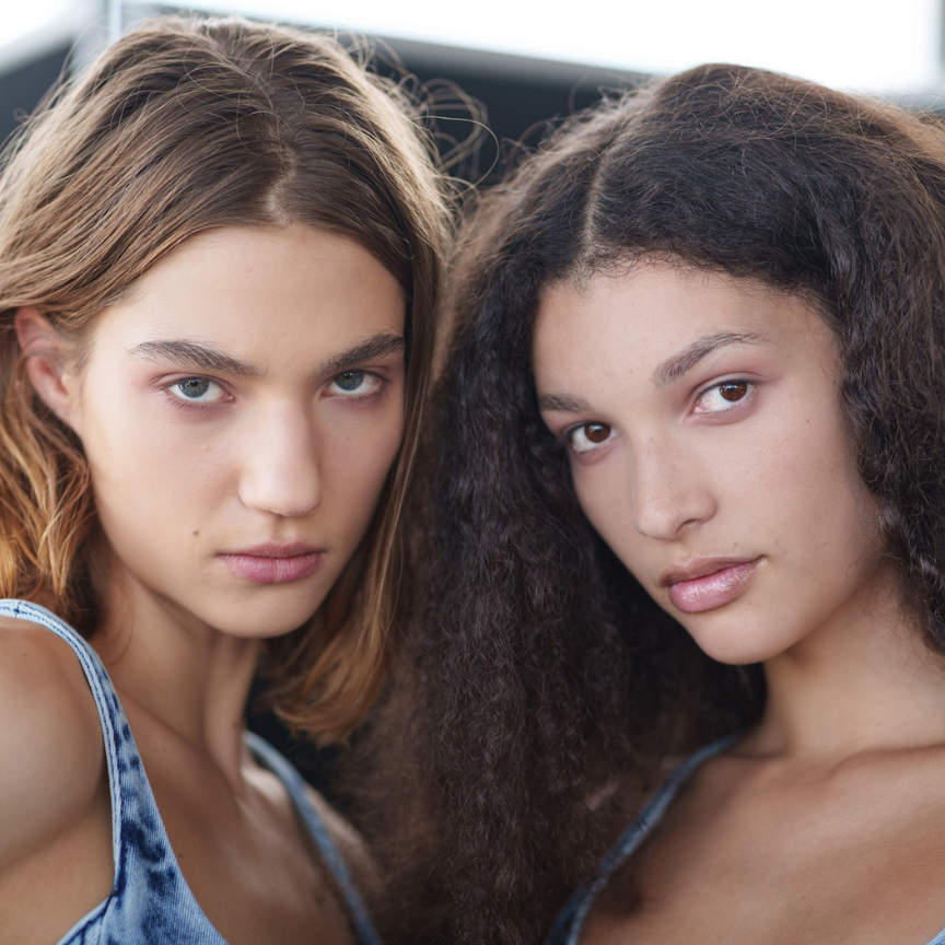 two women facing the camera with bare glowing skin wearing light blue tank tops