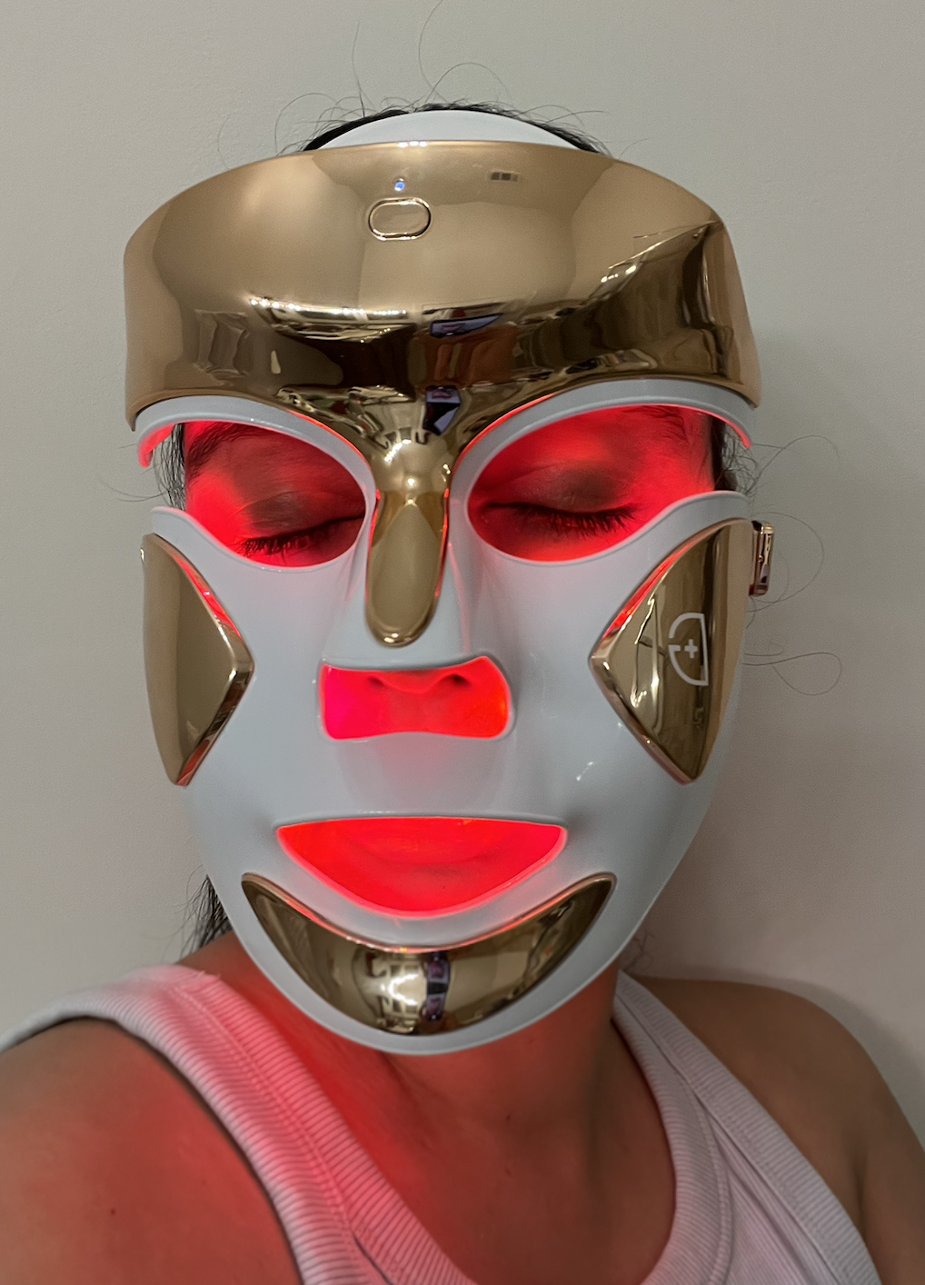 a woman wearing a chrome LED mask with red light therapy and a white tank top