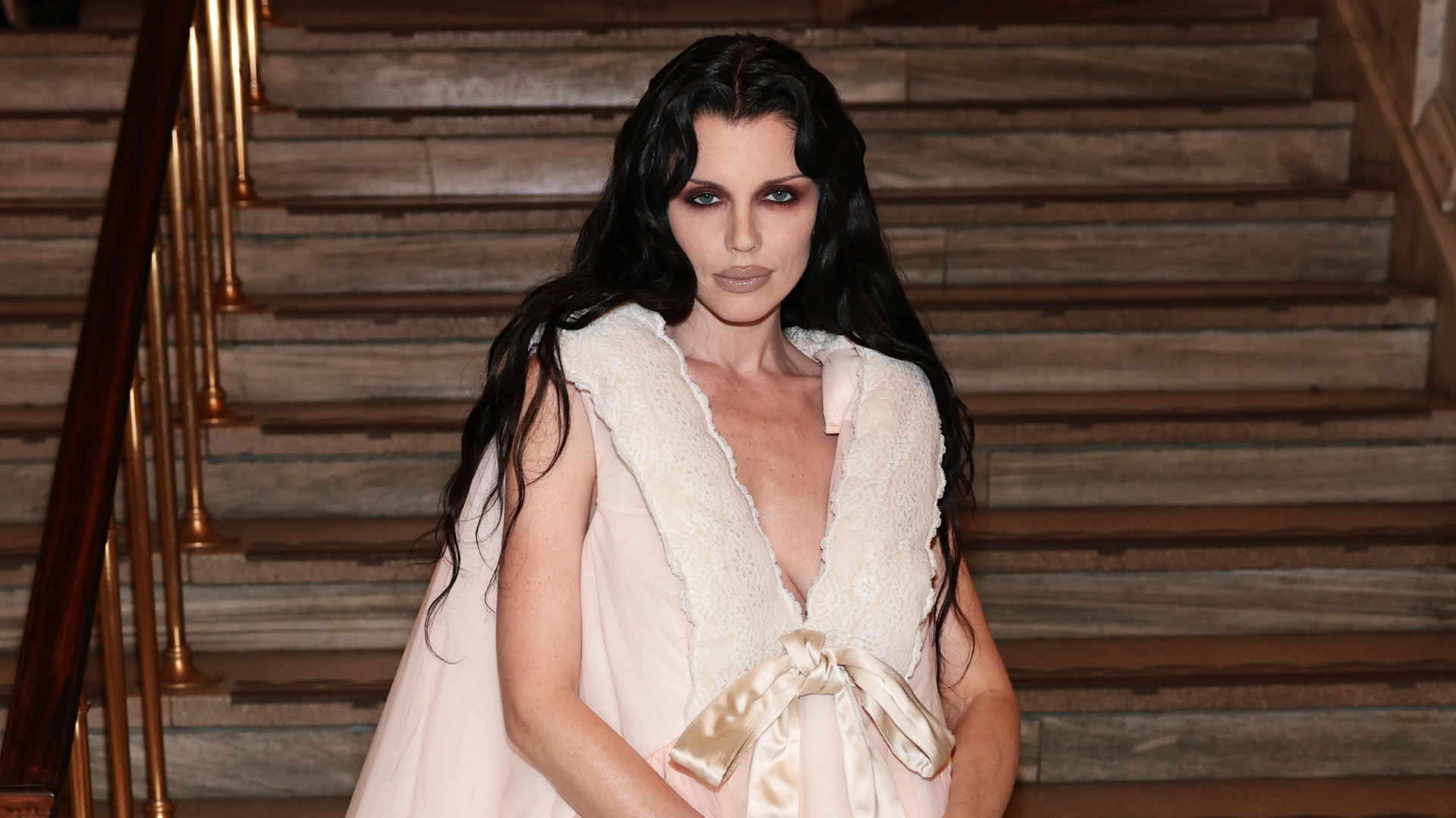 Julia Fox posing in front of steps wearing a frilly dress, dramatic eye makeup and black hair.