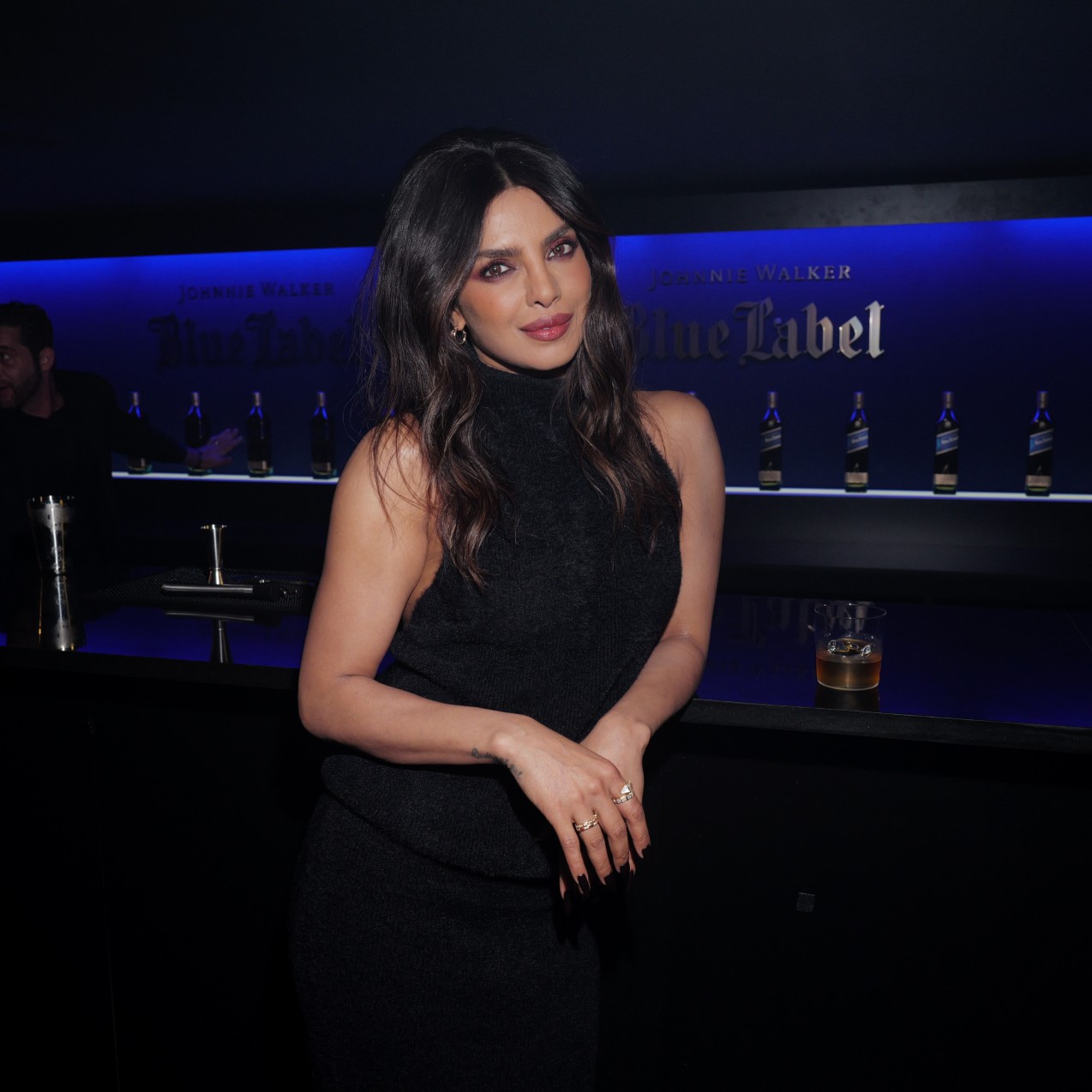 a woman with dark hair wearing a black dress and posing in front of a bar in a dark room