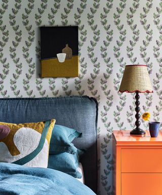 Bedroom with green floral wallpaper, orange side table, lamp, blue bed