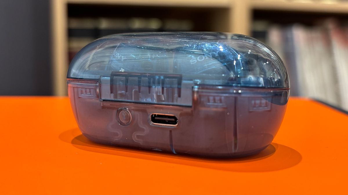 A rear view of the case for a pair of Sony WF-C710N in-ear headphones. It is made of transparent blue plastic and positioned on an orange surface. The USB-C charging port is visible just off centre.