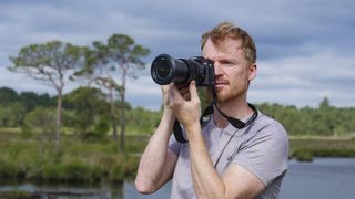 Photographer and tech journalist Tim Coleman holding the Sony RX10 V bridge camera up to his eye in a nature reserve, with dramatic clouds behind him