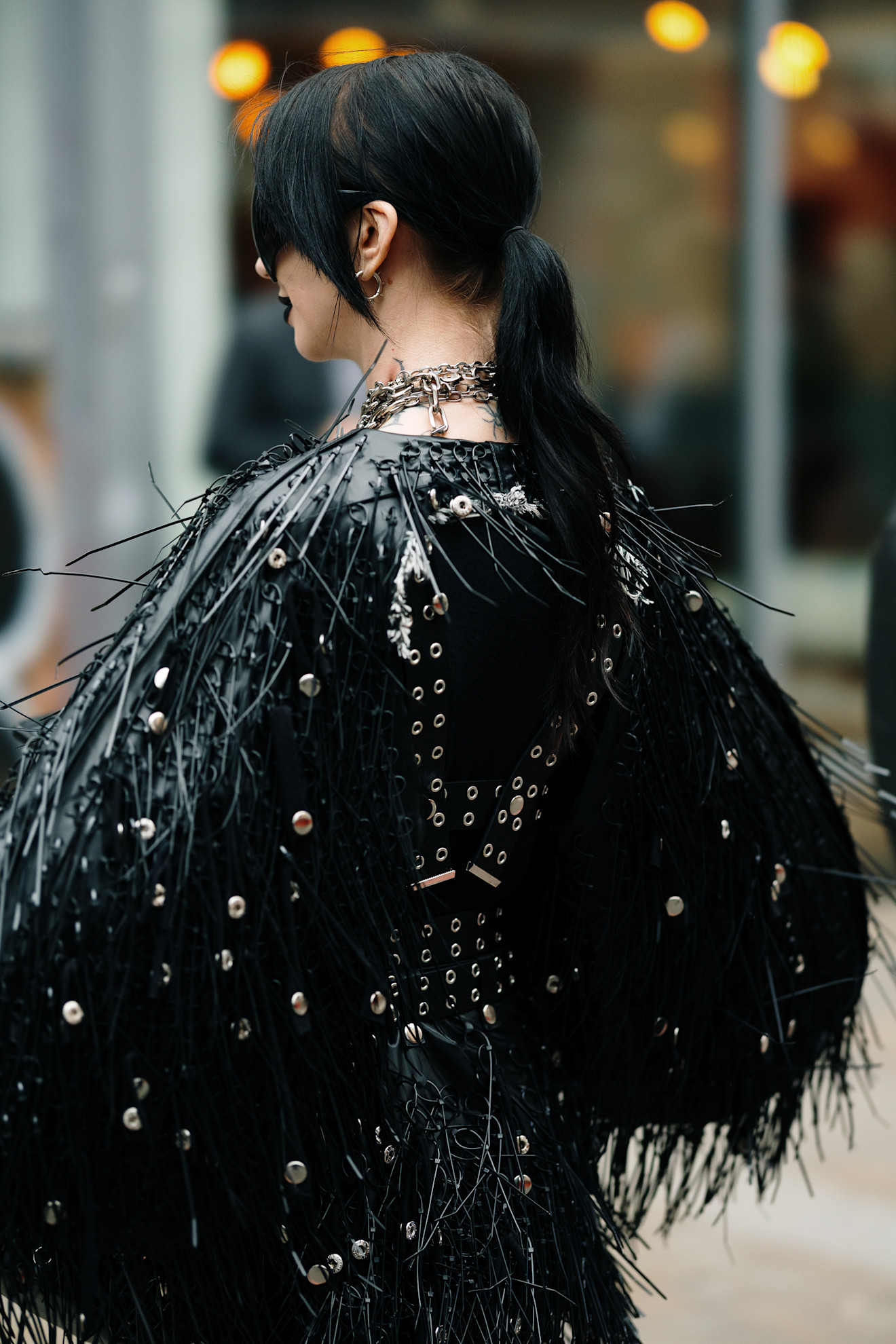 a woman with dark brown hair wearing a black feathered jacket with silver studs and wearing a low ponytail