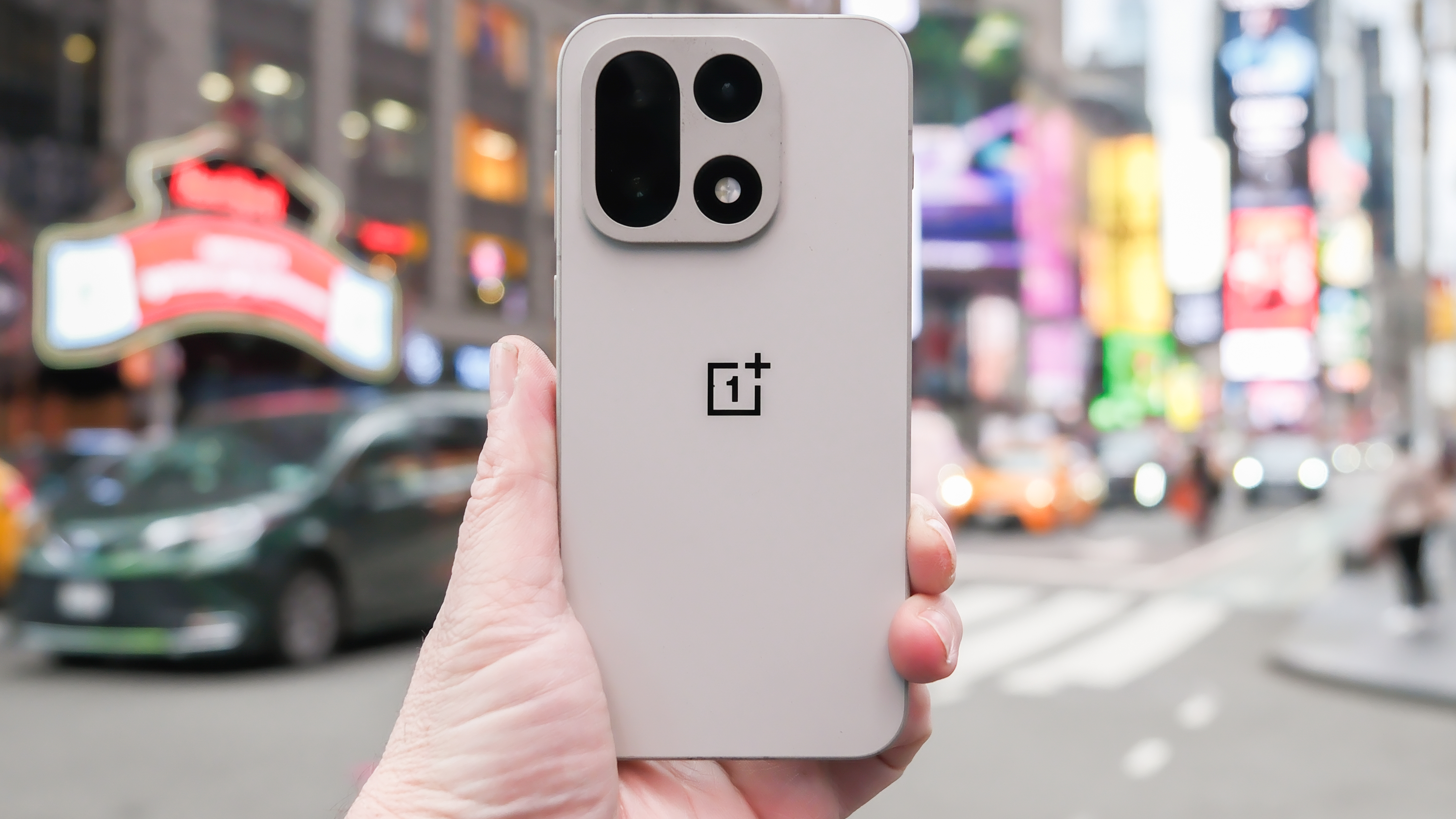 OnePlus 15 in Sand Storm color held in hand with cameras and back showing in the middle of a Times Square New York City intersection across from the Hard Rock Hotel. The background is blurred with cars and people moving past