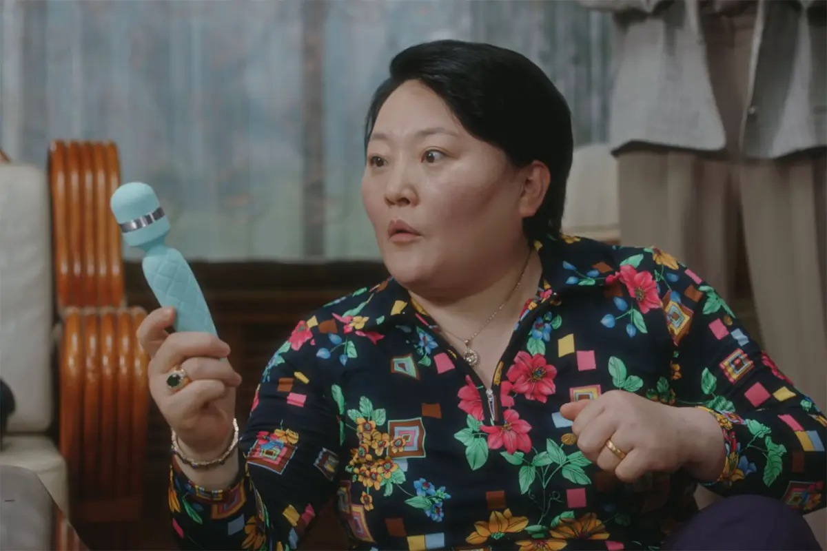 A middle-aged Korean woman with a surprised expression holds a light blue massager, in &#039;A Virtuous Business.&#039;