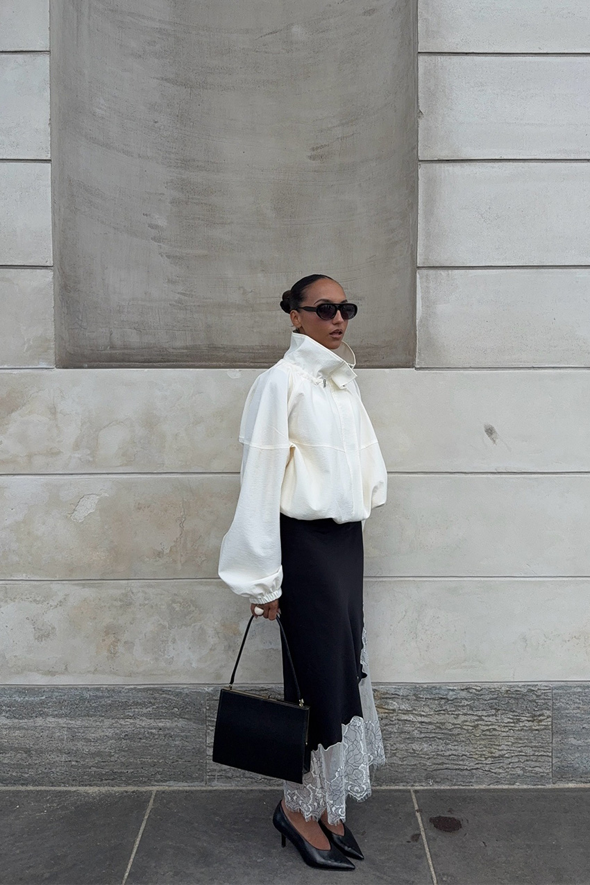 How to wear a slip skirt is shown in a photo of a woman's slip skirt outfit, where she's standing outside in front of a concrete wall, wearing a pair of oversized black aviator sunglasses with a white windbreaker funnel neck jacket styled with a black slip skirt with white lace trim and black pointed pumps and a black frame handbag