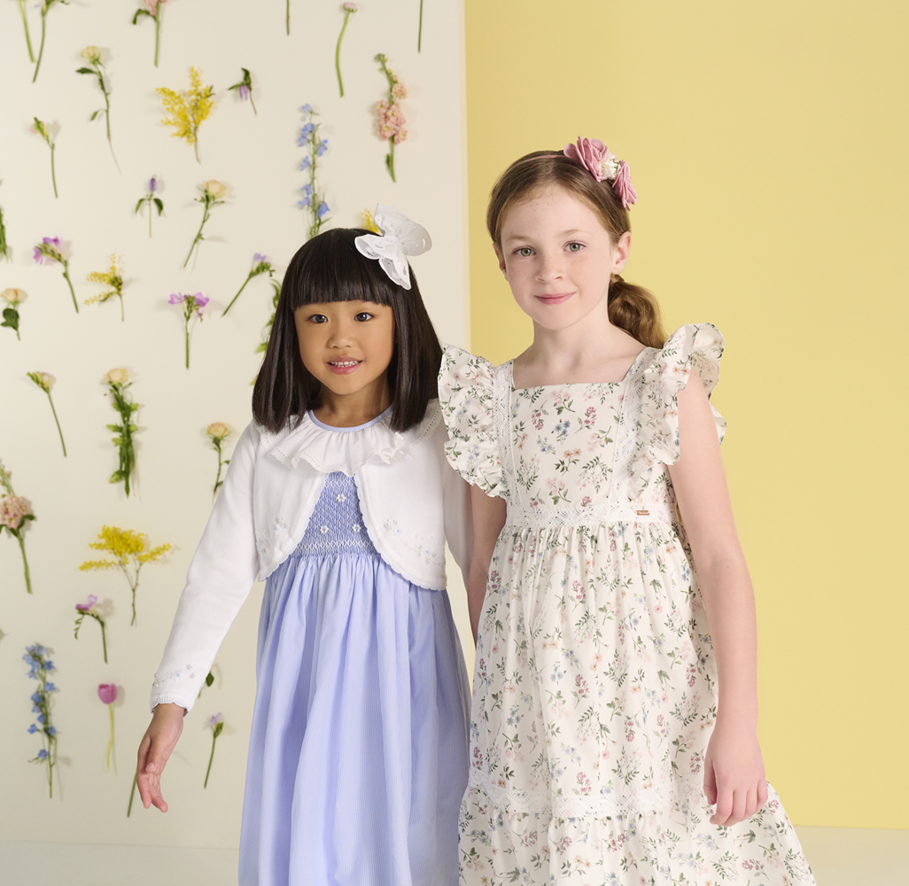 Two girls wearing floral dresses sold at Childrensalon Outlet in front of a yellow wall