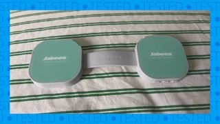 The image shows the flat green and white dumbbell-shaped Jabees PEACE Duo Pillow Speaker on a green and white striped comforter