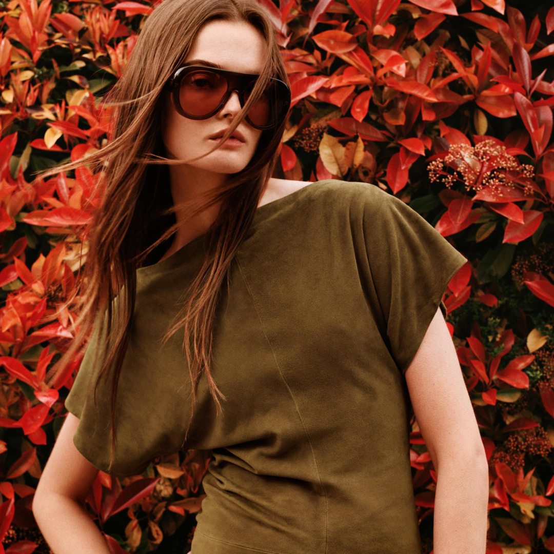 A zara model wearing the Limited Edition Suede Draped Dress and sunglasses standing in front of flowers 